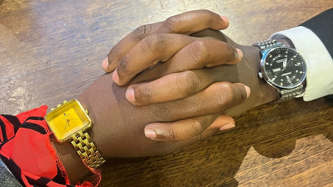 Two clasped hands resting on a wooden table, one wrist wearing a gold square watch, the other a black-dialled steel watch
