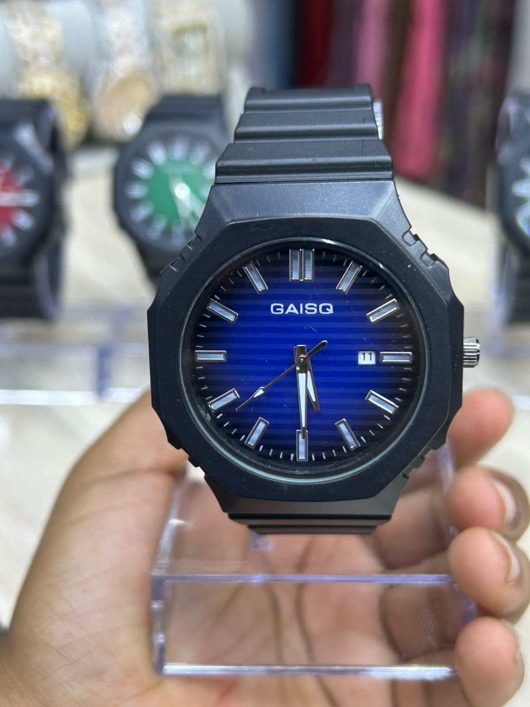 A black octagonal watch with a layered blue dial, held in a hand above the shop counter
